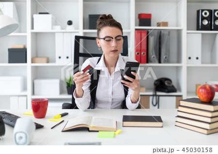 A young girl sits at a computer desk in the office and holds a bank card and phone. 45030158