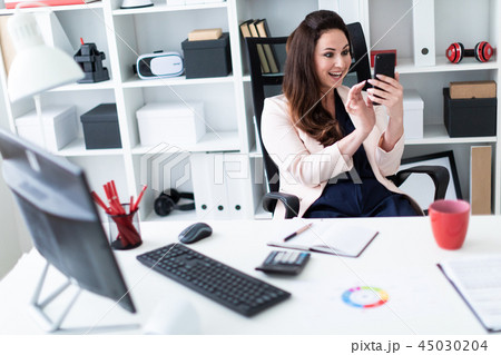 A young girl sitting at a computer Desk in the office, holding a phone and opened her mouth with joy A young girl sitting at a computer Desk in the office, holding a phone and opened her mouth with joy 45030204