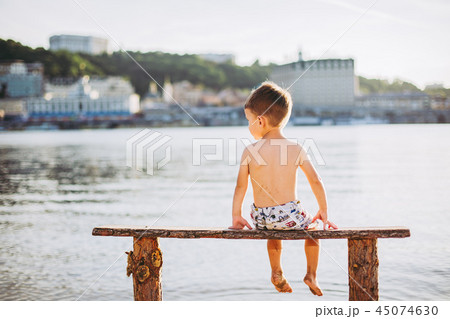 boy child sitting on a wooden bench with his back on the beach near the water and showing his hand boy child sitting on a wooden bench with his back on the beach near the water and showing his hand 45074630