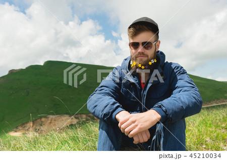 Close-up portrait of a cheerful bearded man in sunglasses and a gray cap with wildflowers in a beard 45210034
