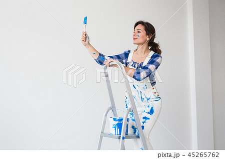 People, renovation and work concept - Beautiful woman standing on the ladder and painting walls 45265762