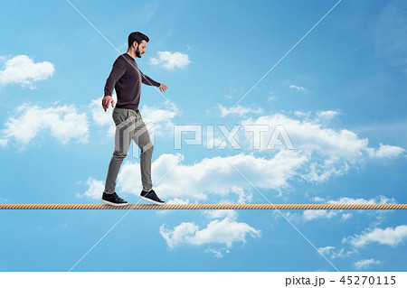 A casually dressed man with a beard walks carefully on a tight rope on a sky background. 45270115