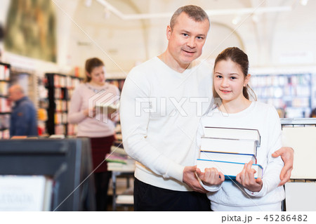 Portrait of tweenage girl with father standing in library with pile of books in hands Portrait of tweenage girl with father standing in library with pile of books in hands 45286482