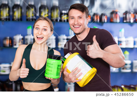 Young muscular people giving thumps up on background with shelves of sport nutrition products 45286650