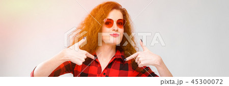 Portrait of beautiful redhead happy young woman in sunglasses smiling showing on blank area on gray Portrait of beautiful redhead happy young woman in sunglasses smiling showing on blank area on gray 45300072