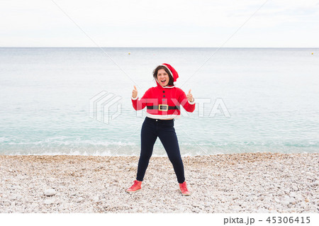 Christmas, humor and people concept - young happy woman in Santa Claus suit at the beach showing 45306415