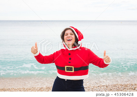 Christmas, humor and people concept - young happy woman in Santa Claus suit at the beach showing 45306428