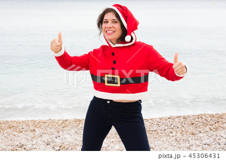 Christmas, humor and people concept - young happy woman in Santa Claus suit at the beach showing 45306431