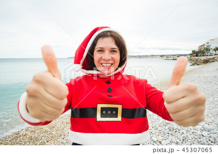 Christmas, humor and people concept - young happy woman in Santa Claus suit at the beach showing Christmas, humor and people concept - young happy woman in Santa Claus suit at the beach showing 45307065