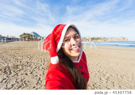 People, holidays and christmas concept - young woman in santa costume taking selfie on beach People, holidays and christmas concept - young woman in santa costume taking selfie on beach 45307756
