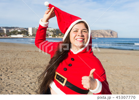 Christmas and holiday concept - Happy woman in Santa Claus costume showing thumbs up on sandy beach Christmas and holiday concept - Happy woman in Santa Claus costume showing thumbs up on sandy beach 45307770