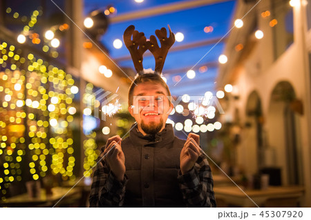 Christmas and holiday concept - Happy man in Christmas deer costume with sparkler Christmas and holiday concept - Happy man in Christmas deer costume with sparkler 45307920