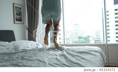 Happy young woman jumps on bed at home having fun in the bed in a luxury apartment with city view in Happy young woman jumps on bed at home having fun in the bed in a luxury apartment with city view in 45324371