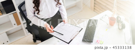 A beautiful young girl stands near an office desk and holds in her hands a sheet for notes and a 45333138