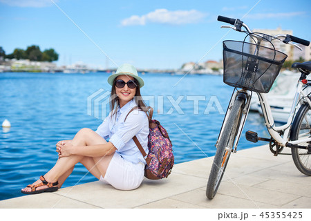 Happy attractive girl in sunglasses and with backpack riding a bike along stony sidewalk Happy attractive girl in sunglasses and with backpack riding a bike along stony sidewalk 45355425