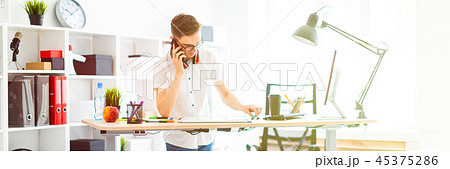 A young man in glasses stands near a computer desk and is talking on the phone. Before him lies a 45375286