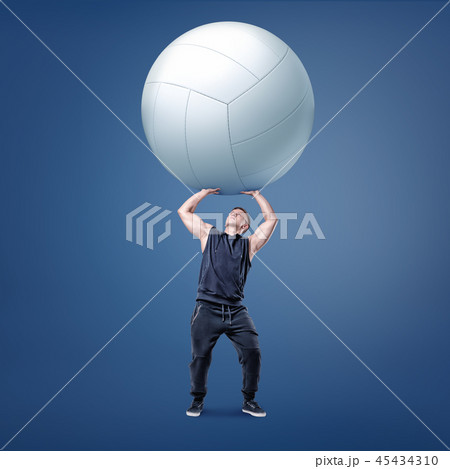 A small muscular athlete holds a large white volleyball ball on straight arms above himself. A small muscular athlete holds a large white volleyball ball on straight arms above himself. 45434310