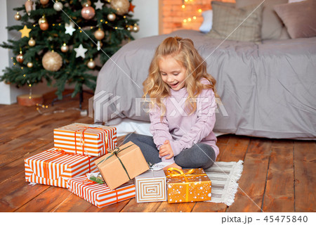 Christmas morning, a girl in pajamas with blond hair is sitting near the bed and looking at the Christmas morning, a girl in pajamas with blond hair is sitting near the bed and looking at the 45475840