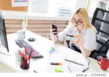 A young girl sitting in the office and holding a phone and a Bank card. 45508360