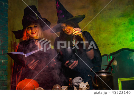 Portrait of smiling two witches in black hats reading book at table with pumpkin and skulls 45522140