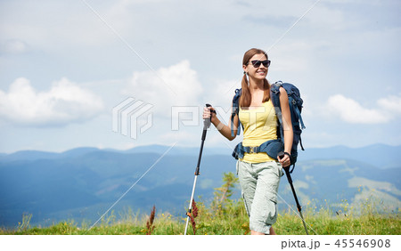 Woman hiker hiking on grassy hill, wearing backpack, using trekking sticks in the mountains 45546908