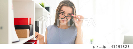 Beautiful young girl in an office stands near a shelf with documents and holds her hand glasses. Beautiful young girl in an office stands near a shelf with documents and holds her hand glasses. 45595323