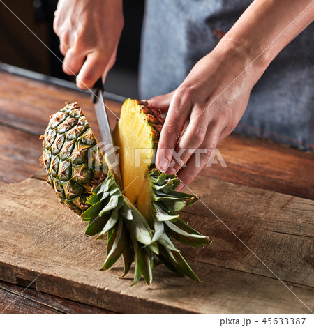 The girl's hand with a knife cuts a tropical fruit pineapple on a board on a wooden brown table The girl's hand with a knife cuts a tropical fruit pineapple on a board on a wooden brown table 45633387