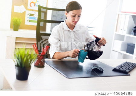 A young girl sitting at a table in the office and pours coffee into a Cup. A young girl sitting at a table in the office and pours coffee into a Cup. 45664299