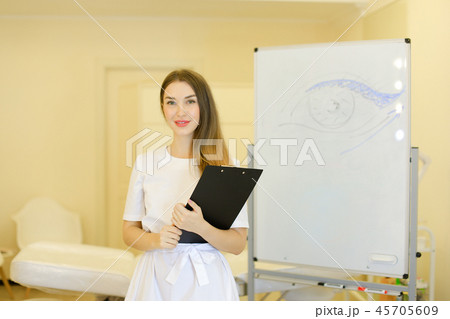 Young cosmetologist standing with black folder near placard with drawn eye at cosmetology cabinet. 45705609