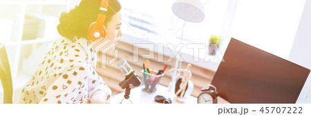 Beautiful young girl sits in headphones and with a notepad at the table in the office and speaks Beautiful young girl sits in headphones and with a notepad at the table in the office and speaks 45707222