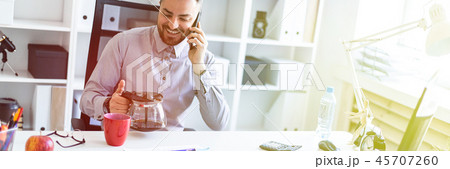A young man in the office sits at a table, talking on the phone and holding a coffee pot. 45707260