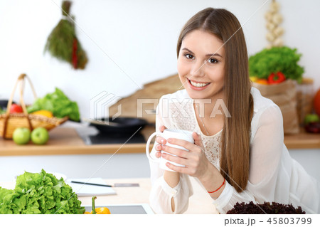 Young happy woman holding white cup and looking at the camera while sitting at wooden table in the Young happy woman holding white cup and looking at the camera while sitting at wooden table in the 45803799