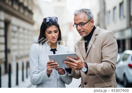 Man and woman business partners with tablet standing outdoors in city, talking. 45807400