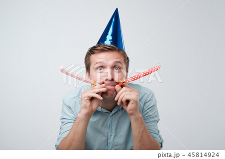 cheerful young man having fun on party wearing blue denim shirt and holiday hat, blowing party horn. 45814924