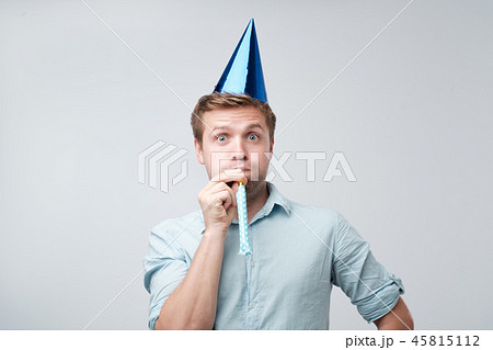 cheerful young man having fun on party wearing blue denim shirt and holiday hat, blowing party horn. 45815112