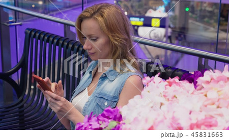 A beautiful young woman is sitting on a bench in a large modern shopping center near a flower bed A beautiful young woman is sitting on a bench in a large modern shopping center near a flower bed 45831663