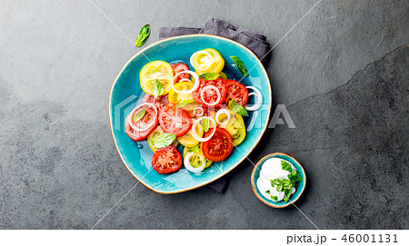 Red and yellow fresh tomato salad with onion, basil and white sauce on blue plate. Gray slate Red and yellow fresh tomato salad with onion, basil and white sauce on blue plate. Gray slate 46001131