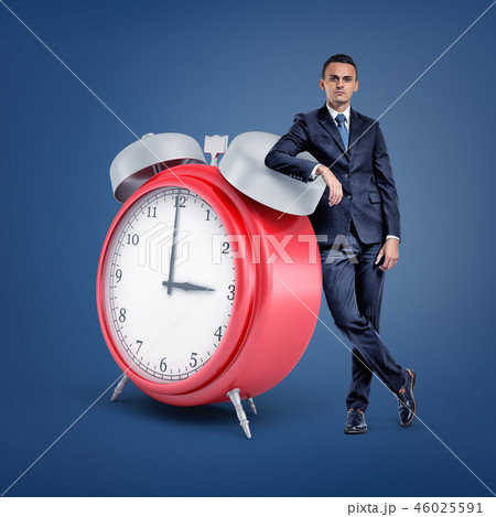 A small businessman stands looking at front and leaning on a huge red alarm clock. 46025591