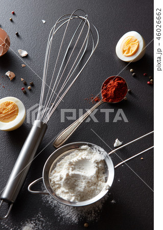 Sieve with flour, spoon with red pepper, a metal whisk and halves of a boiled egg on a black Sieve with flour, spoon with red pepper, a metal whisk and halves of a boiled egg on a black 46026662