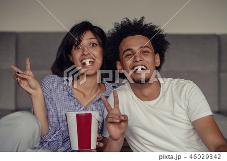 Image of smiling couple sitting on floor near sofa at home and looking aside while eating popcorn Image of smiling couple sitting on floor near sofa at home and looking aside while eating popcorn 46029342