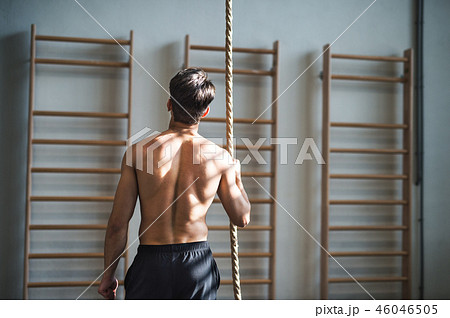 Fit young man in gym standing topless , holding a climbing rope. Rear view. Fit young man in gym standing topless , holding a climbing rope. Rear view. 46046505