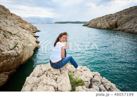 Back view of young smiling woman in jeans and T-shirt sitting alone on stony shore of sea Back view of young smiling woman in jeans and T-shirt sitting alone on stony shore of sea 46057199