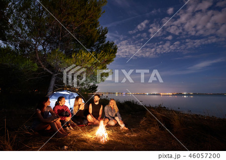 Night summer camping on shore. Group of young tourists around campfire near tent under evening sky Night summer camping on shore. Group of young tourists around campfire near tent under evening sky 46057200