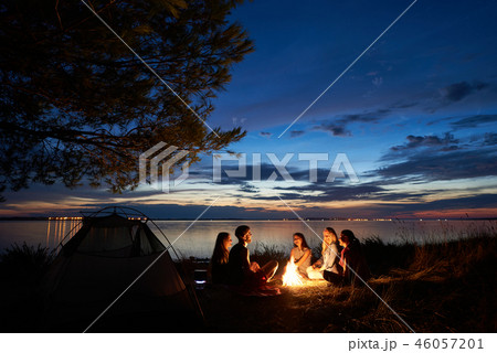 Night summer camping on shore. Group of young tourists around campfire near tent under evening sky 46057201
