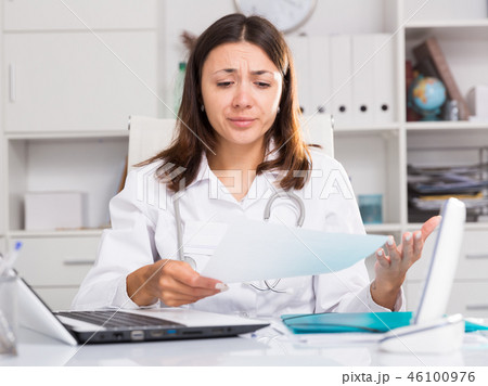 Upset girl doctor in uniform with papers at laptop in hospital Upset girl doctor in uniform with papers at laptop in hospital 46100976