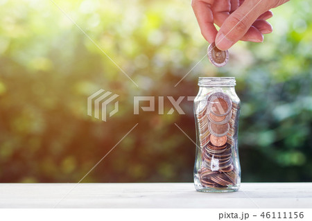 Hand holding coin over coins in glass jar. 46111156
