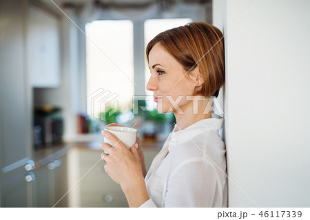 A young woman with cup of coffee standing indoors, leaning on a wall. 46117339