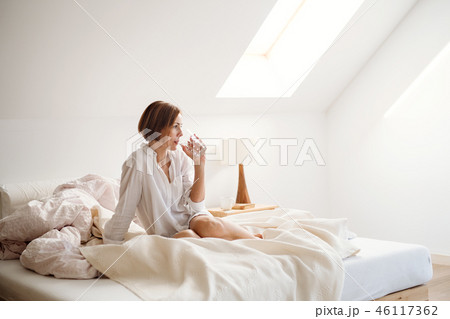 A young woman with night shirt sitting indoors on bed in the morning, drinking water. A young woman with night shirt sitting indoors on bed in the morning, drinking water. 46117362