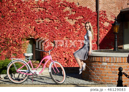 Attractive blond girl at pink lady bicycle on sunny day on wall overgrown with red ivy background. 46129083