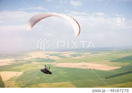 Professional paraglider in a cocoon suit flies high above the ground against the sky and fields 46167270